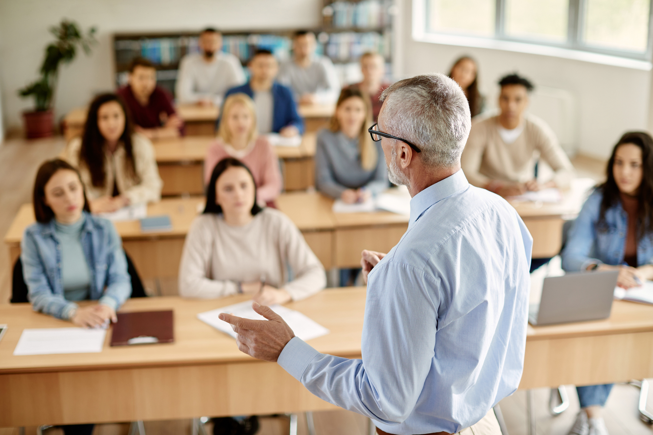 A man with white hair and a light blue shirt stands and speaks in front of a class of young adults seated at rows of wooden desks. Earn your adult education certificate online from the University of San Diego.