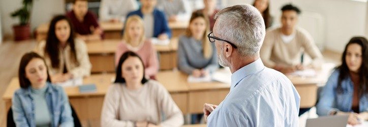 The image shows a classroom scene where an older male teacher, facing away from the camera, is addressing a group of diverse, attentive students. The students, variously engaged and looking towards the teacher, are seated in a typical lecture hall setting. This setting highlights an interactive and inclusive educational environment. The image effectively captures a moment of active learning and could be used to discuss various teaching philosophies that emphasize engagement and communication between instructors and students.