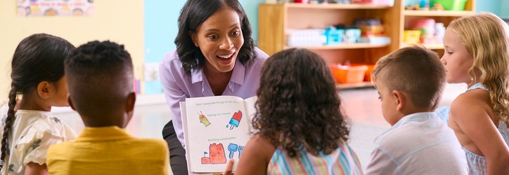 a teacher with dark hair holds out a book to a group of student surrounding her on the floor, leading a reading session