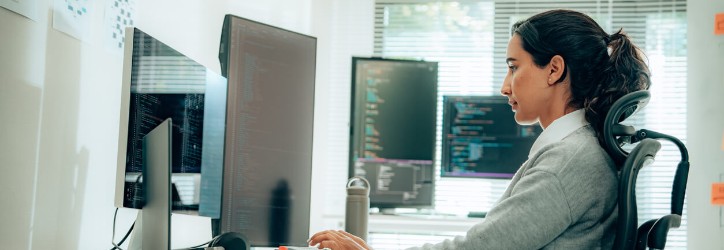 An adult Hispanic woman focused on coding on dual monitors, exhibiting dedication and concentration in a modern, well-lit office environment.