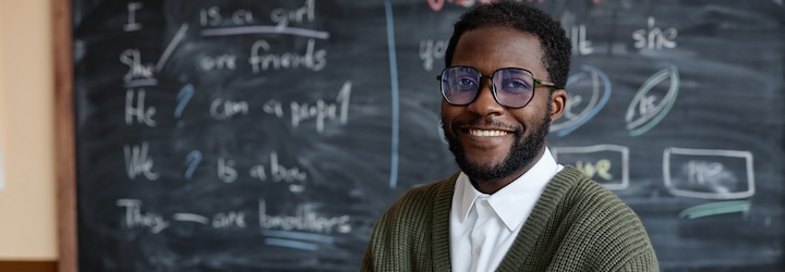 Medium portrait of young African American male teacher of English wearing eyeglasses sitting at desk in classroom smiling at camera while using laptop.