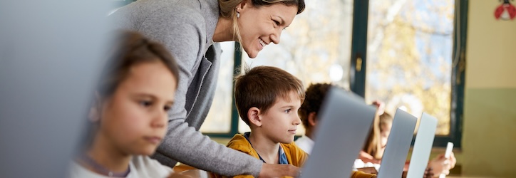 Happy female teacher assisting school kids during an e-learning class at elementary school. Focus is on boy.