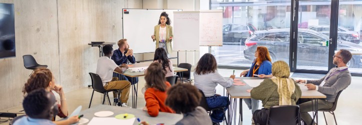A class of adults listening to a woman speak in front of a whiteboard.