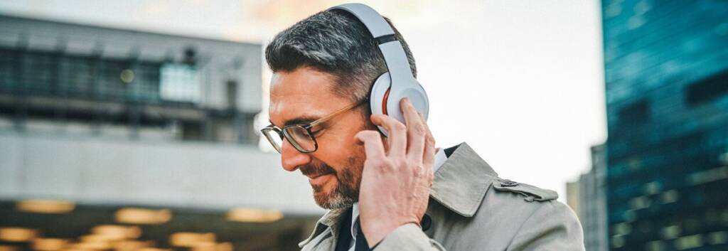 A smiling man in glasses and a beige trench coat adjusts his white over-ear headphones while looking at a smartphone, with modern city buildings behind him